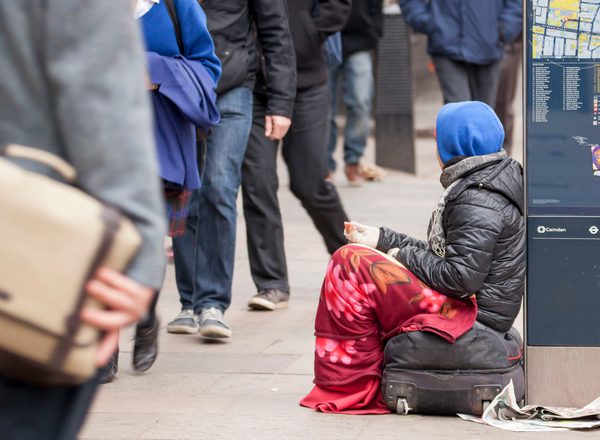 Female beggar in London. Sitting on a bag, with a blanket round her legs and a blue hood and brown coat. A newspaper and litter is on the path as she rests with a hand out against a map as people walk past