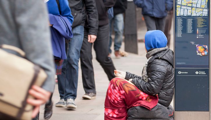 Female beggar in London. Sitting on a bag, with a blanket round her legs and a blue hood and brown coat. A newspaper and litter is on the path as she rests with a hand out against a map as people walk past