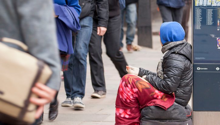 Female beggar in London. Sitting on a bag, with a blanket round her legs and a blue hood and brown coat. A newspaper and litter is on the path as she rests with a hand out against a map as people walk past
