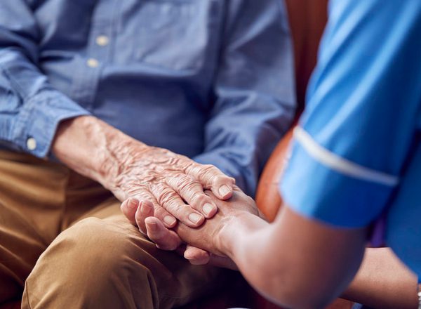 Close Up Of Female Care Worker In Uniform Holding Hands Of Senior Man Sitting In Care Home Lounge