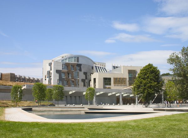 The Scottish parliament building rises in the backdrop, foregrounded by a pond, trees and a lawn. Blue skies overhead