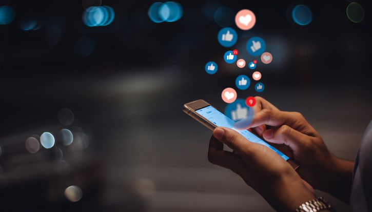 Someone's hands holding a mobile phone, with social media likes and heart icons floating out of it like bubbles