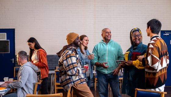 Adults standing in a circle and talking to each other about their mental wellbeing. There is a staff member sitting to the left of them in the frame sitting next to a table. The community centre is located in Seaton Deleval in the North East of England.
