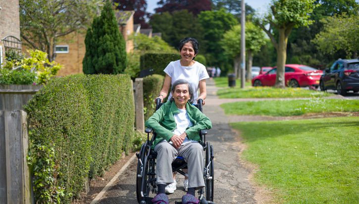 A female carer pushes an older female wheelchair user along a path in a residential area on a sunny day