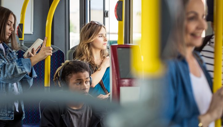 The inside of a bus. Lots of people sit or stand on a bus during daylight