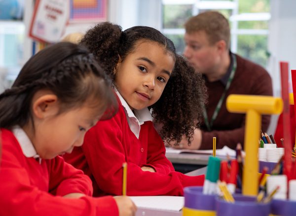 A little girl in a red school uniform looks at the camera while another student writes with a pencil next to her