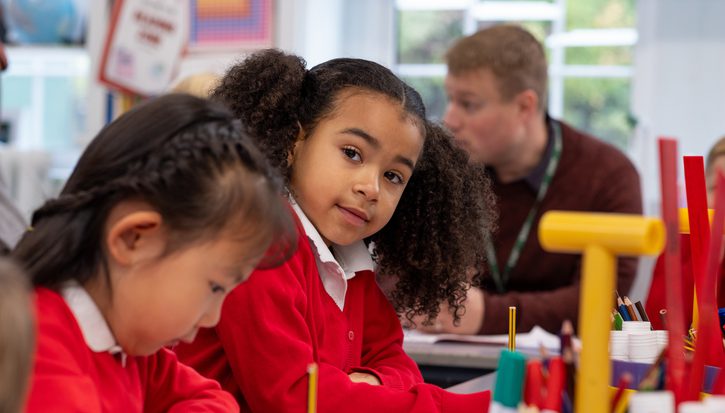 A little girl in a red school uniform looks at the camera while another student writes with a pencil next to her