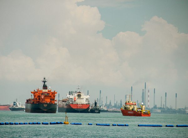 A wide shot of several large commercial vessels, including oil tankers and a survey boat, floating on turquoise water. In the background, an industrial shoreline with numerous smokestacks and refinery infrastructure is visible under a cloudy sky. Concept of global logistics, maritime industry, and international trade.