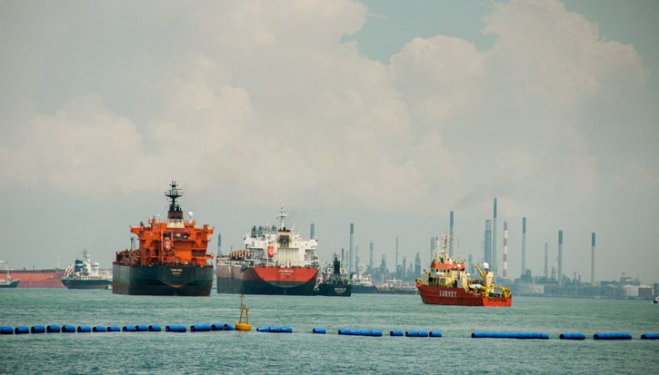 A wide shot of several large commercial vessels, including oil tankers and a survey boat, floating on turquoise water. In the background, an industrial shoreline with numerous smokestacks and refinery infrastructure is visible under a cloudy sky. Concept of global logistics, maritime industry, and international trade.