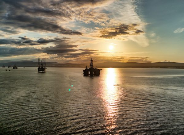 Dramatic high-angle drone photograph of decommissioned oil platforms anchored in the Cromarty Firth, near Invergordon, Scotland, as the sun sets over a calm North Sea