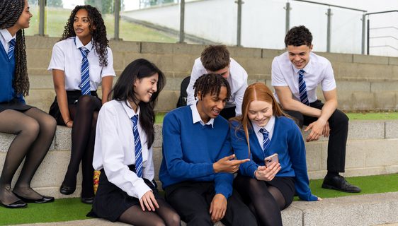 A group of young people wearing school uniforms sit on some concrete steps looking at a phone screen and chatting