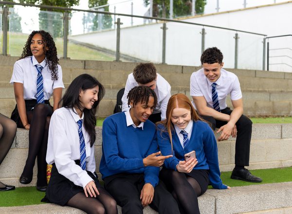 A group of young people wearing school uniforms sit on some concrete steps looking at a phone screen and chatting