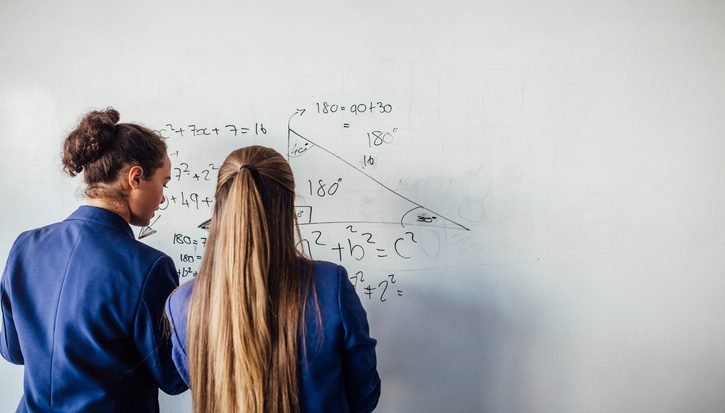 Two teenage girls can be seen from behind wearing blue school blazers working out a maths equation on a whiteboard