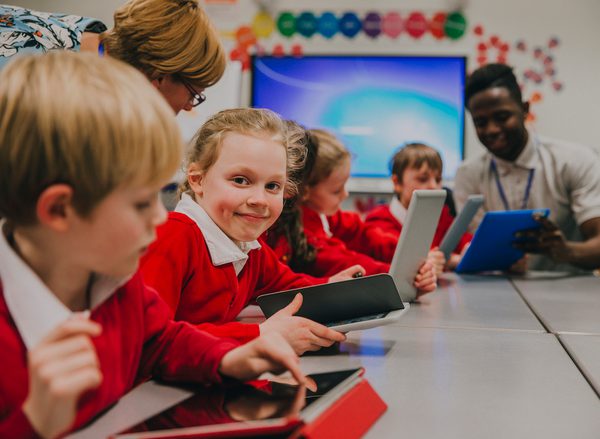 A group of primary school aged children using ipads around a table, a little girl smiles at the camera