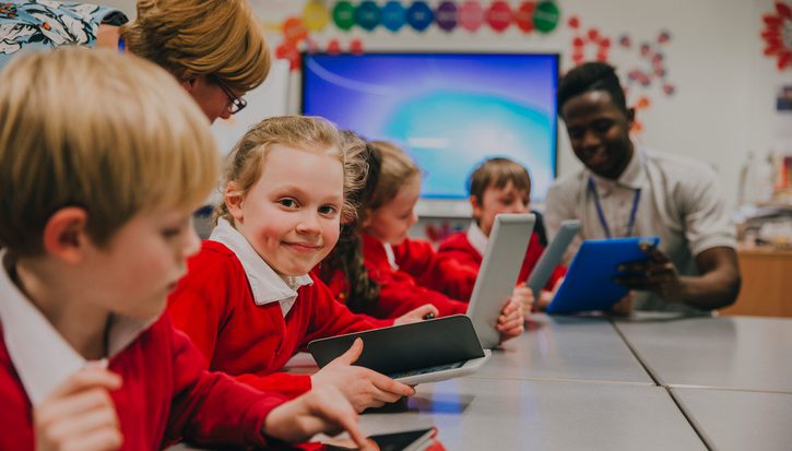 A group of primary school aged children using ipads around a table, a little girl smiles at the camera