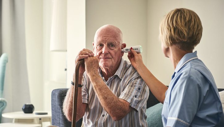 An older man with a walking stick is sitting on a sofa having his temperature taken with a thermometer in his ear by a female nurse in uniform