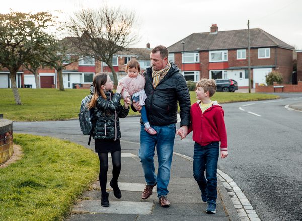 A photo of a family walking along a residential street: a dad holds a toddler in his arms with a son and daughter on either side of him
