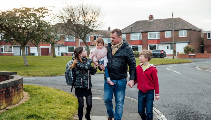 A photo of a family walking along a residential street: a dad holds a toddler in his arms with a son and daughter on either side of him