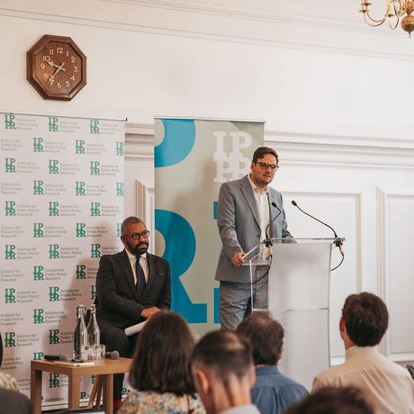 Harry Quilter-Pinner is stood at a lectern speaking. James Cleverly is sat to the left.