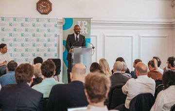 Audience facing James Cleverly, who is stood at a lectern speaking.