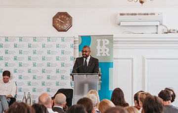 Audience facing James Cleverly, who is stood at a lectern speaking.
