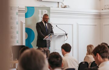 Audience facing James Cleverly, who is stood at a lectern speaking.