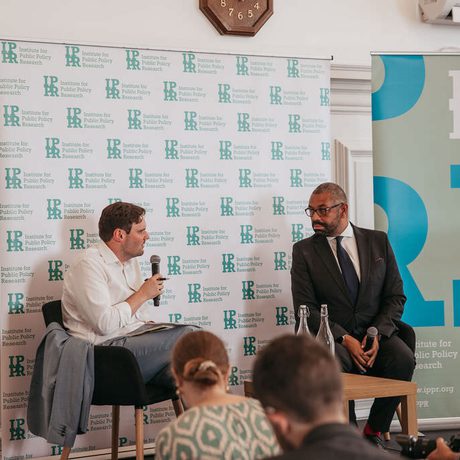 James Cleverly and Harry Quilter-Pinner sat in front of IPPR banners. Harry Quilter-Pinner is speaking into a microphone.